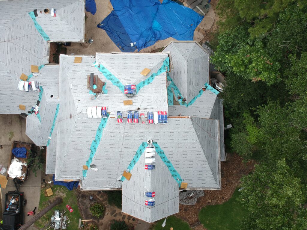 Picture of a roof from above, stripped of shingles, with new felt underlayment and the ice/water shield laid down on the plywood decking. The pool in back is covered by tarps. 